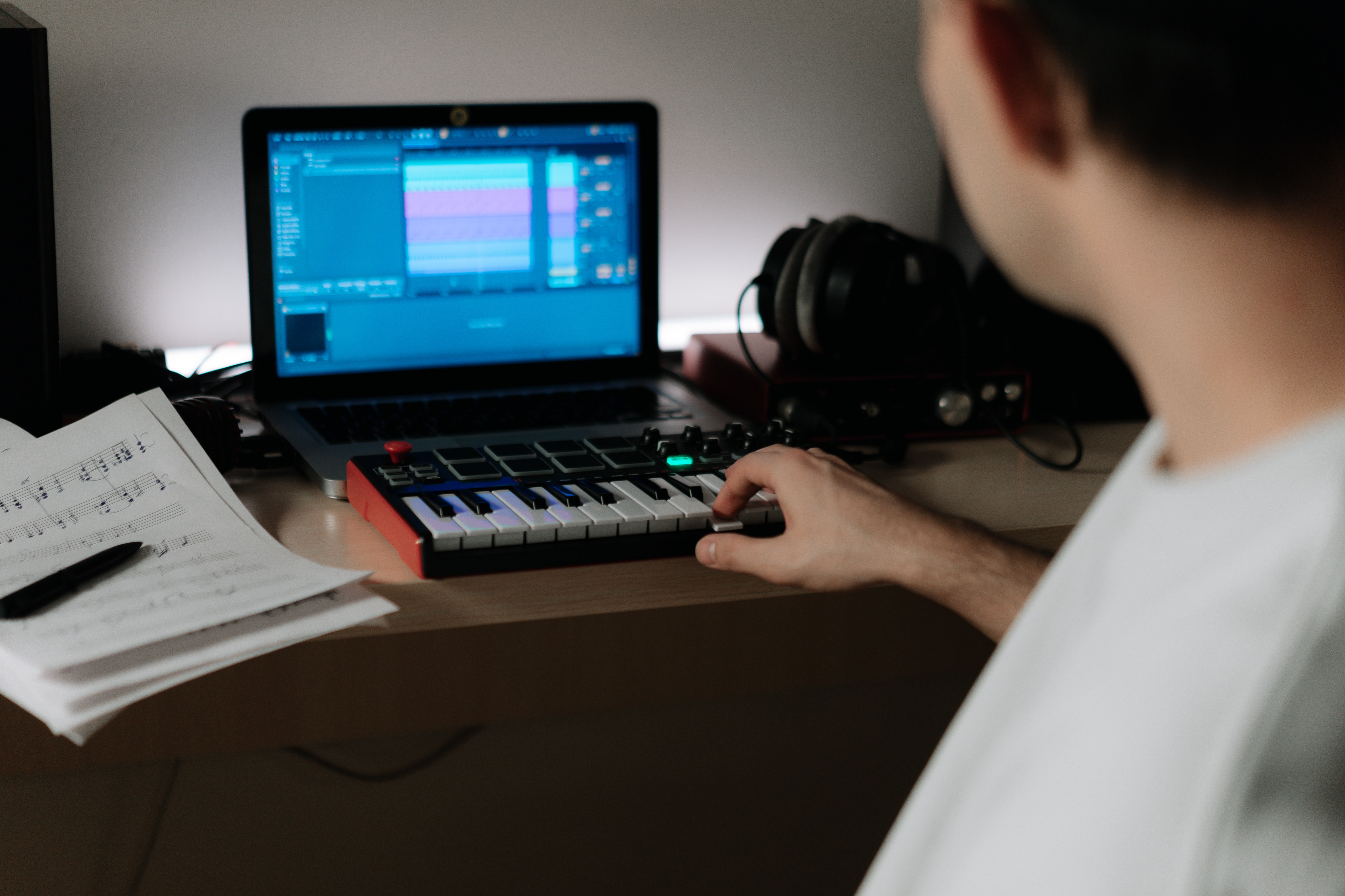 Person producing a song with a midi keyboard, a laptop, and a music sheet nearby